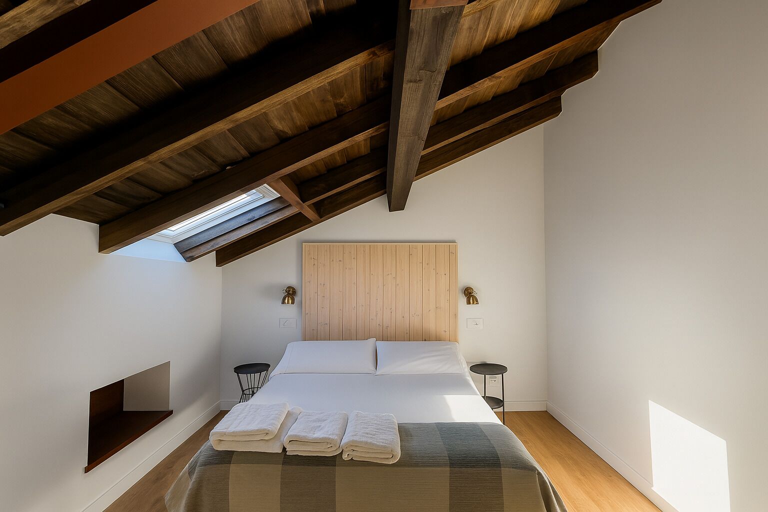 Un dormitorio con techo inclinado, una cama con ropa de cama blanca, dos mesitas de noche y un tragaluz.