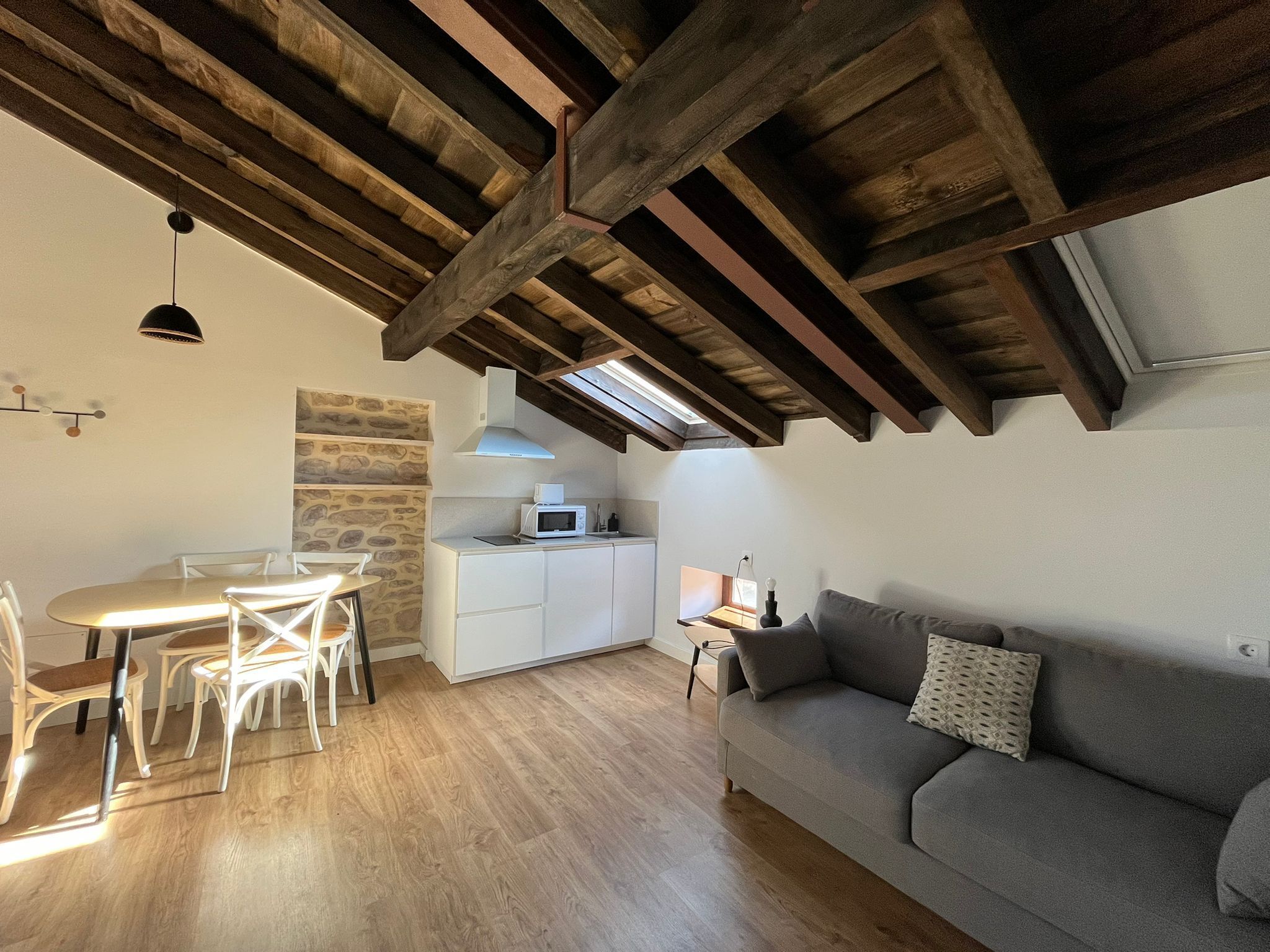 A cozy loft-style room with exposed wooden beams, a dining area with a table and chairs, a kitchenette with a microwave, and a gray sofa with a patterned pillow.