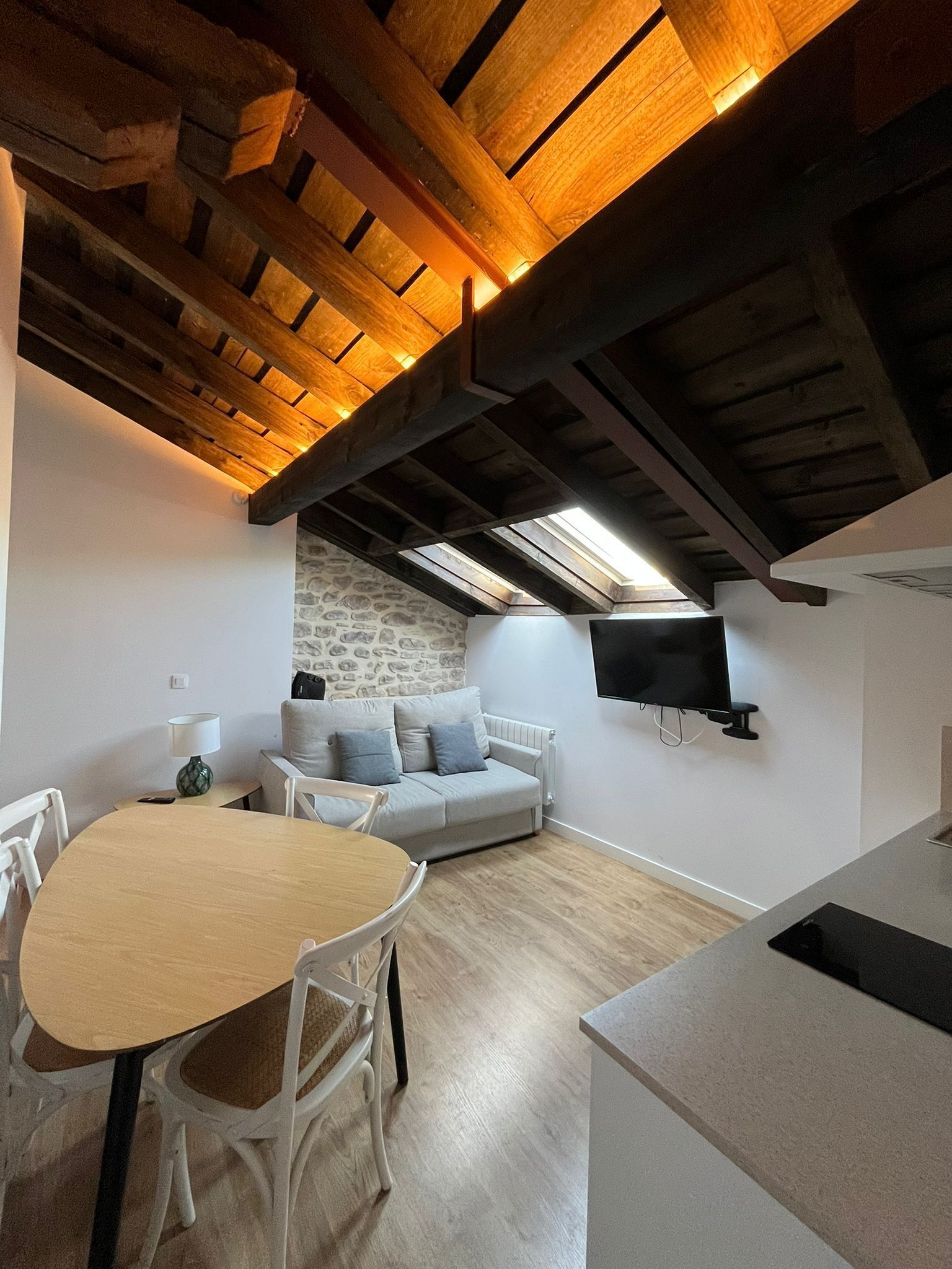 Um quarto de sótão aconchegante com teto de madeira, parede de pedra, sofá cinza, mesa de jantar e televisão de tela plana fixada na parede.