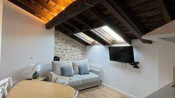 Um quarto de sótão aconchegante com teto de madeira, parede de pedra, sofá cinza, mesa de jantar e televisão de tela plana fixada na parede.