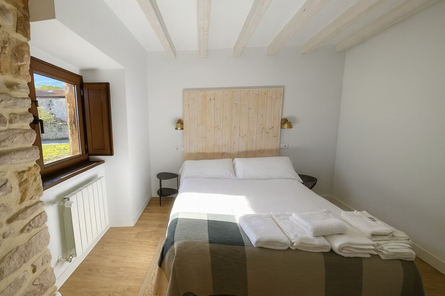 Una camera da letto con una testiera in legno, un letto con lenzuola bianche, un comodino e una finestra che si affaccia sul verde.