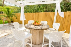 A round wooden table with a fruit bowl, surrounded by white chairs, under a white pergola.