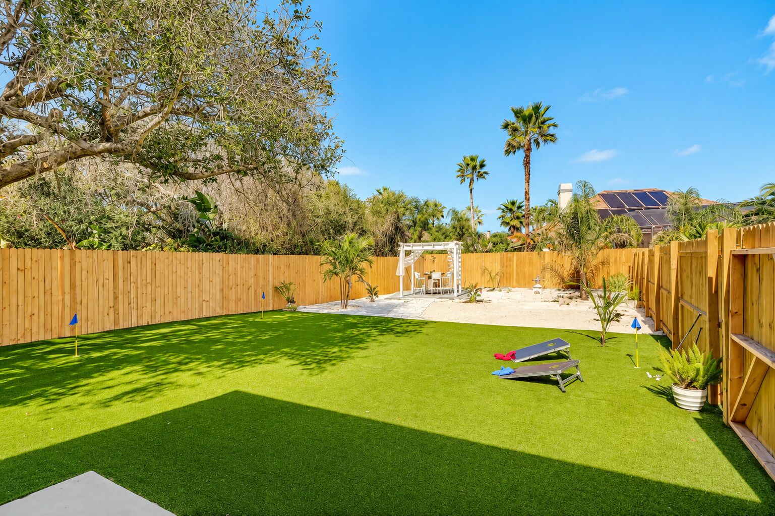 Un jardin avec une pelouse artificielle, une clôture en bois, des palmiers et une pergola.