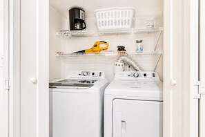 A laundry room with two white Whirlpool washers, a yellow hairdryer, a coffee maker, and a white laundry basket on a shelf.