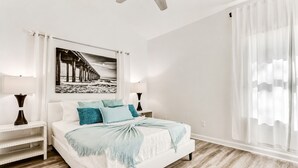 A bedroom with a bed, nightstands, a lamp, a painting, and a window with curtains. - Lux Beach Cabana - Close to Beach & Restaurants (St. Augustine)