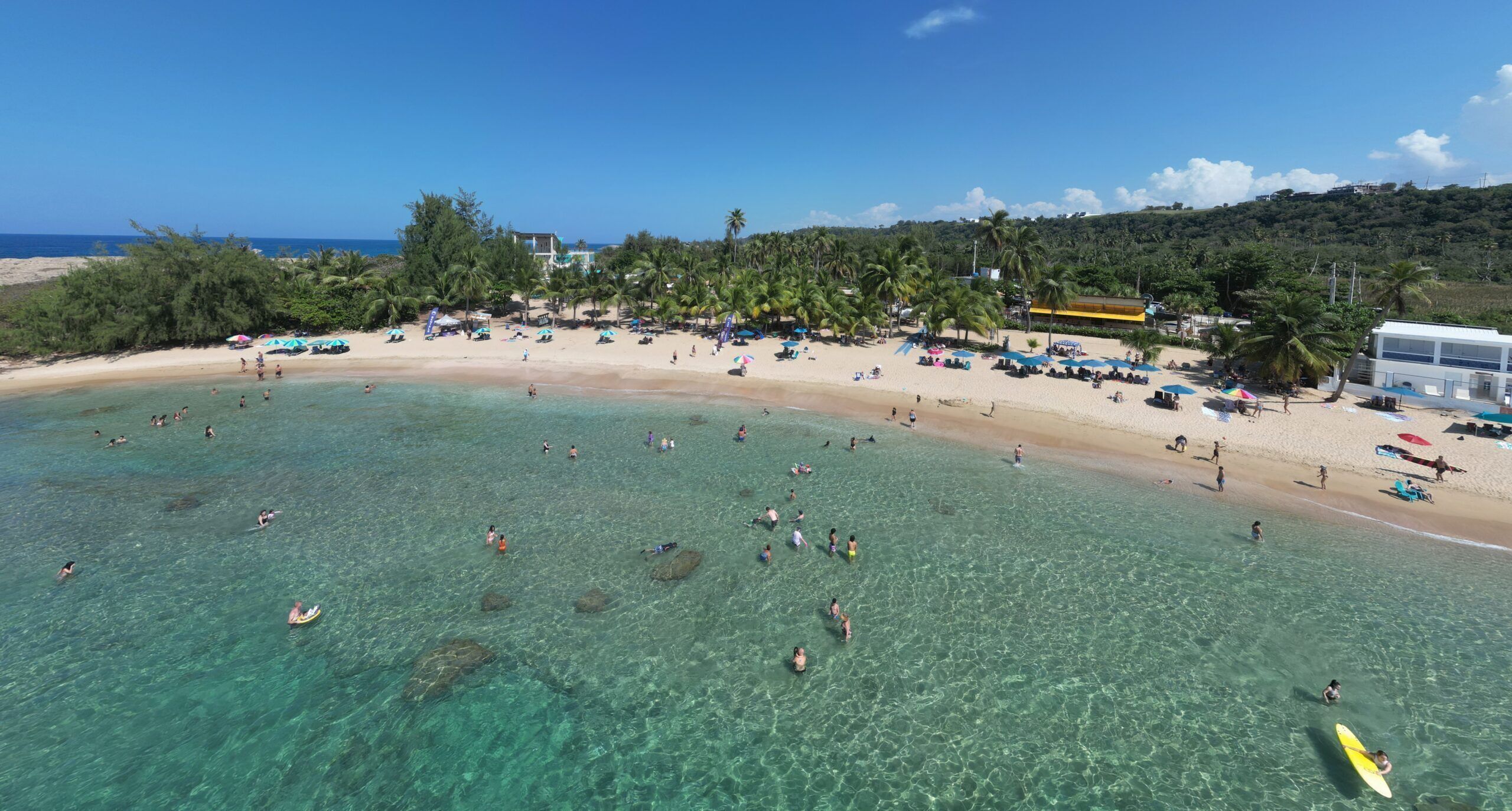 Une plage avec une eau turquoise et limpide, des gens qui se baignent et prennent des bains de soleil, et un bâtiment avec un toit jaune en arrière-plan.