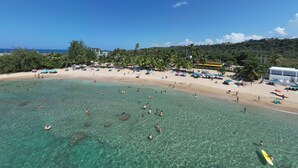 A beach with clear turquoise water, people swimming and sunbathing, and a building with a yellow roof in the background.