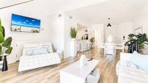 A modern, well-lit living room with a white sofa, a white coffee table, and a large painting of a beach scene.