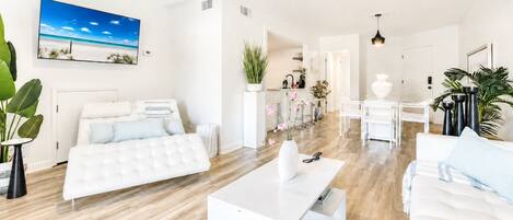 A modern, well-lit living room with a white sofa, a white coffee table, and a large painting of a beach scene.