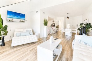 A modern, well-lit living room with a white sofa, a white coffee table, and a large painting of a beach scene.