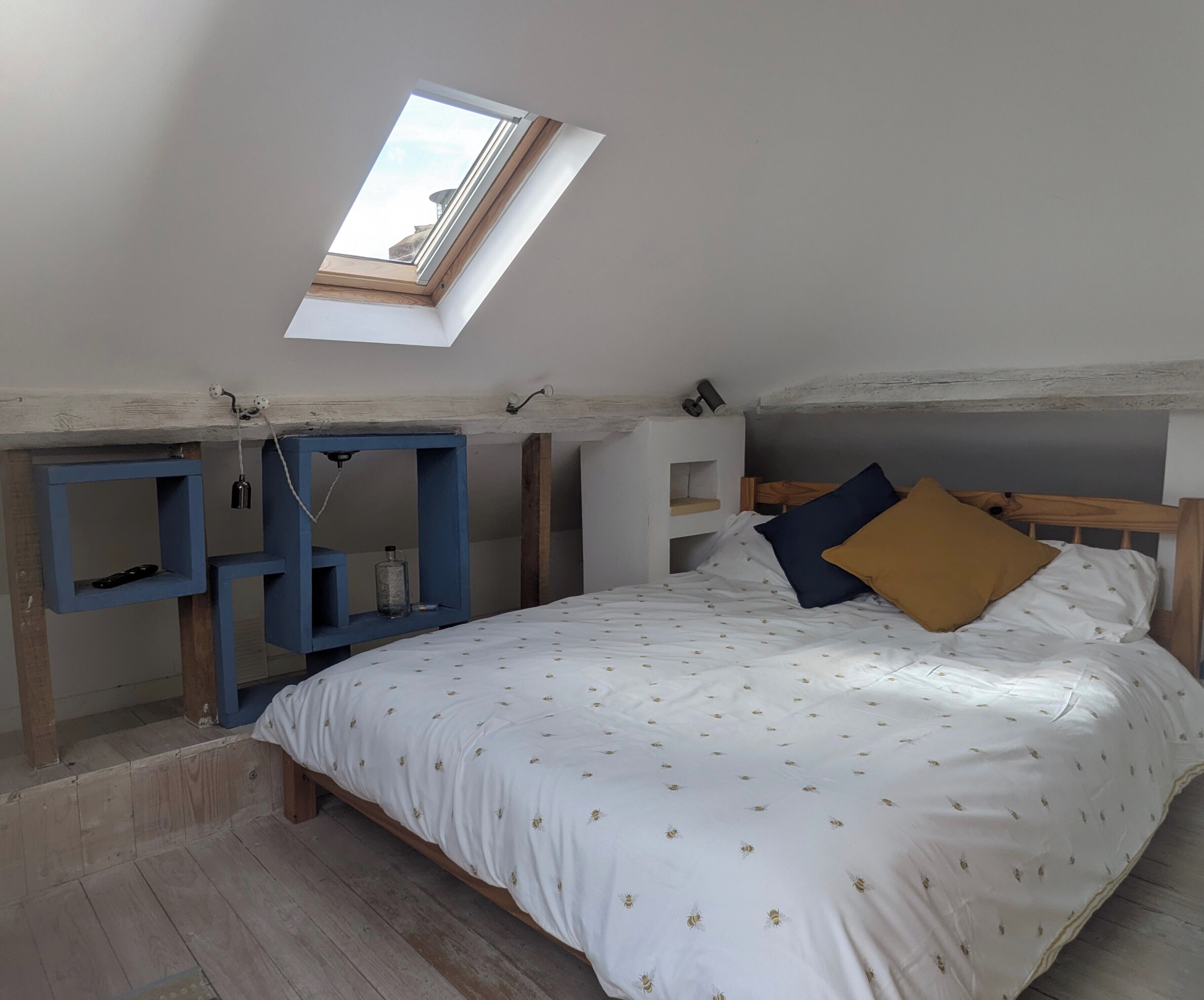 A bedroom with a sloped ceiling, a skylight, a bed with white bedding and patterned pillows, and built-in wooden shelves.