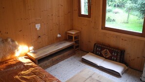 Puinen huone, jossa on sänky, pieni pöytä, penkki ja ikkuna, josta avautuu näkymä vehreään maisemaan.