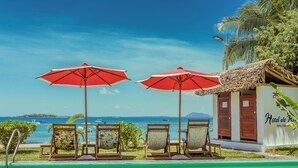 Zona de piscina con sillones de descanso y sombrillas rojas, un edificio pequeño con la leyenda 'Hotel de Rio', y vistas al mar y a islas distantes.