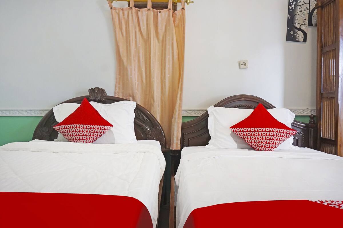 Une chambre avec deux lits, chacun étant équipé de draps blancs et d’oreillers rouges. Il y a un rideau à la fenêtre, et un interrupteur fixé au mur est visible.