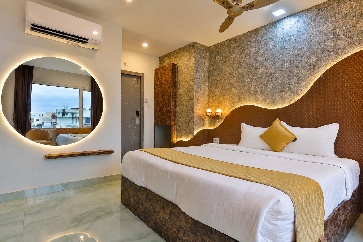A modern hotel room with a large bed, a circular mirror, a ceiling fan, and a view of the cityscape.