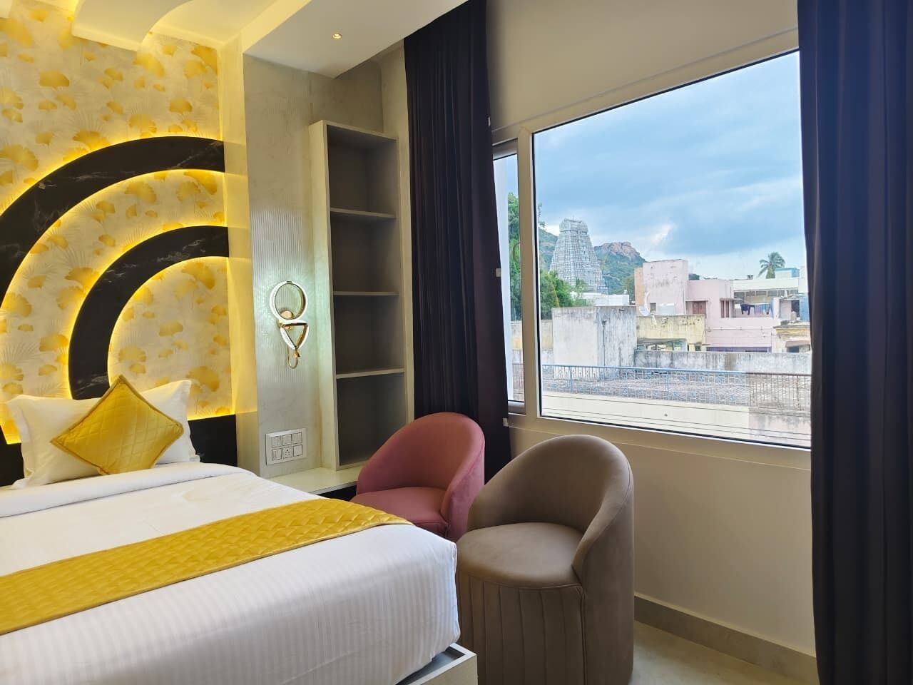 A hotel room with a bed, a chair, a shelf, and a large window with a view of buildings.