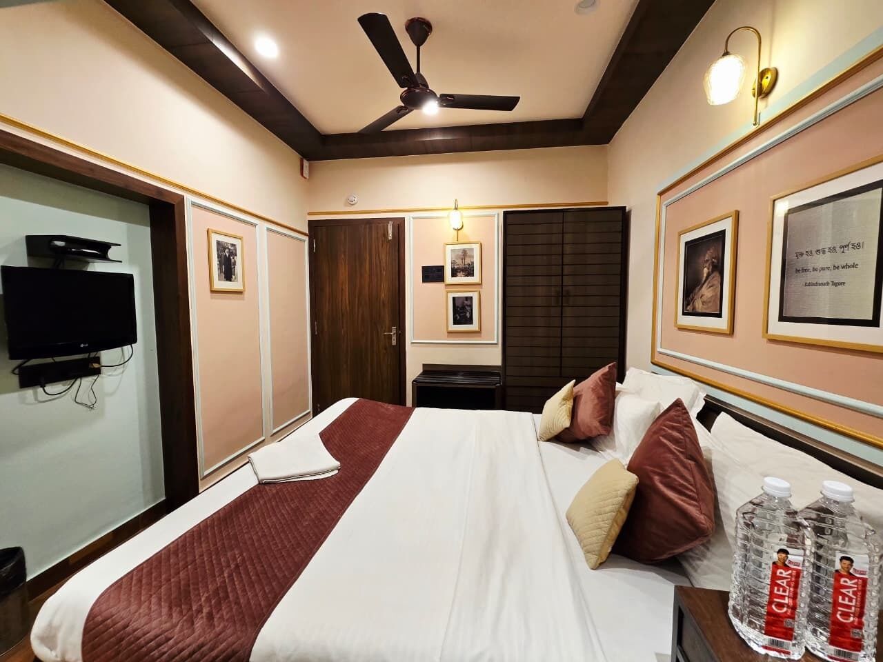 A hotel room with a bed, a ceiling fan, a television, and framed pictures on the walls.