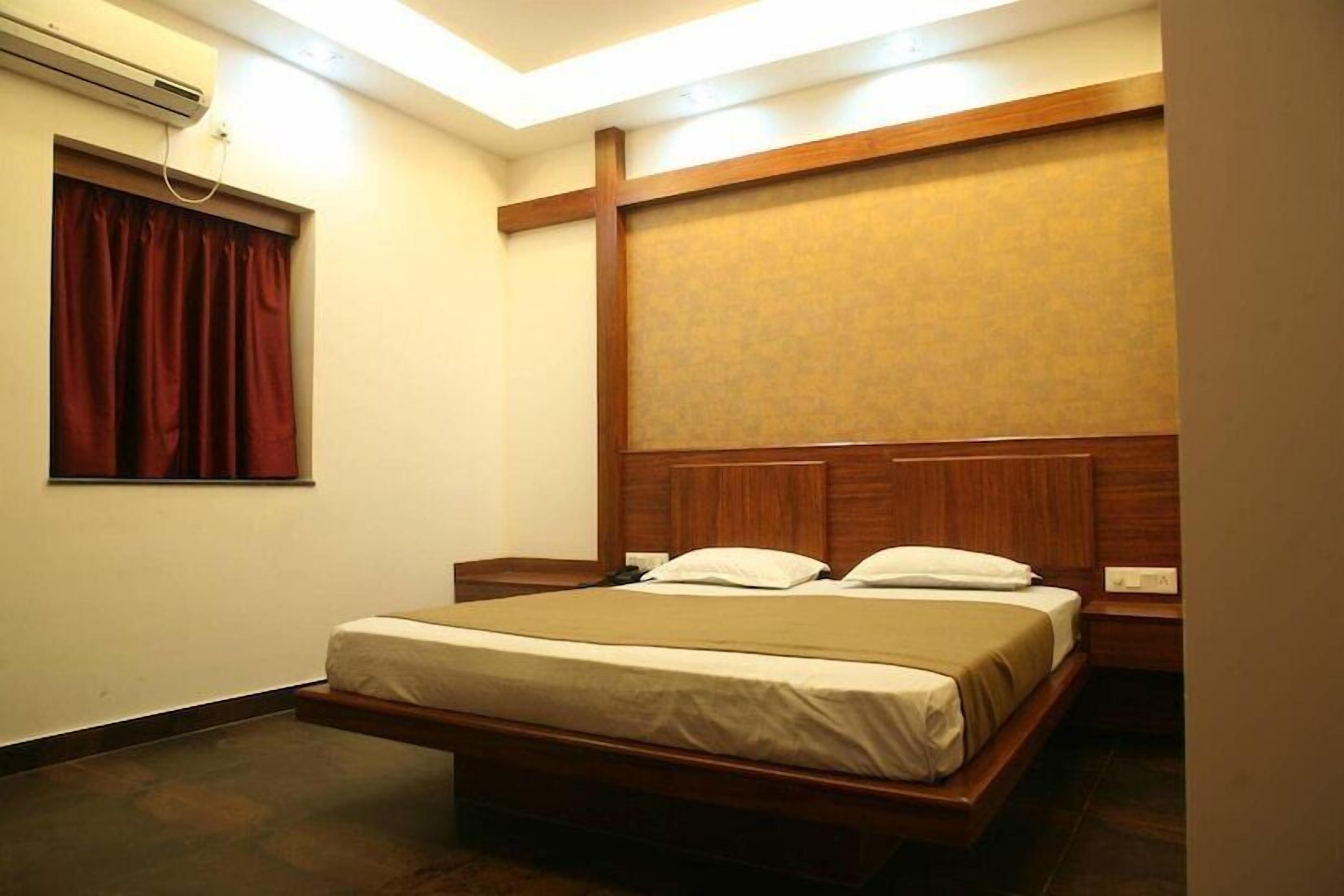 A hotel room with a wooden bed, a small bedside table, a nightstand, a window with red curtains, and an air conditioning unit on the wall.