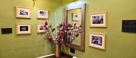 A bathroom with a green wall, framed photographs, a mirror, a vase with flowers, and a wooden vanity with a remote control and cups.
