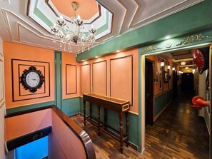A hallway with wooden flooring, a wooden bench, a chandelier, and a clock on the wall. - Hotel Trimoorti (Kolkata)