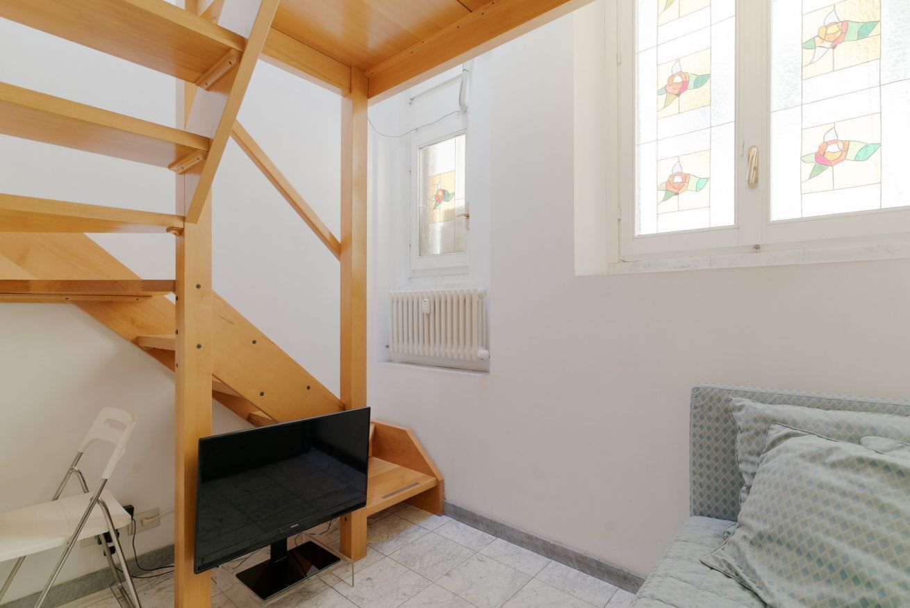 A room with a wooden staircase, a bed, a TV, a radiator, and a window with stained glass.