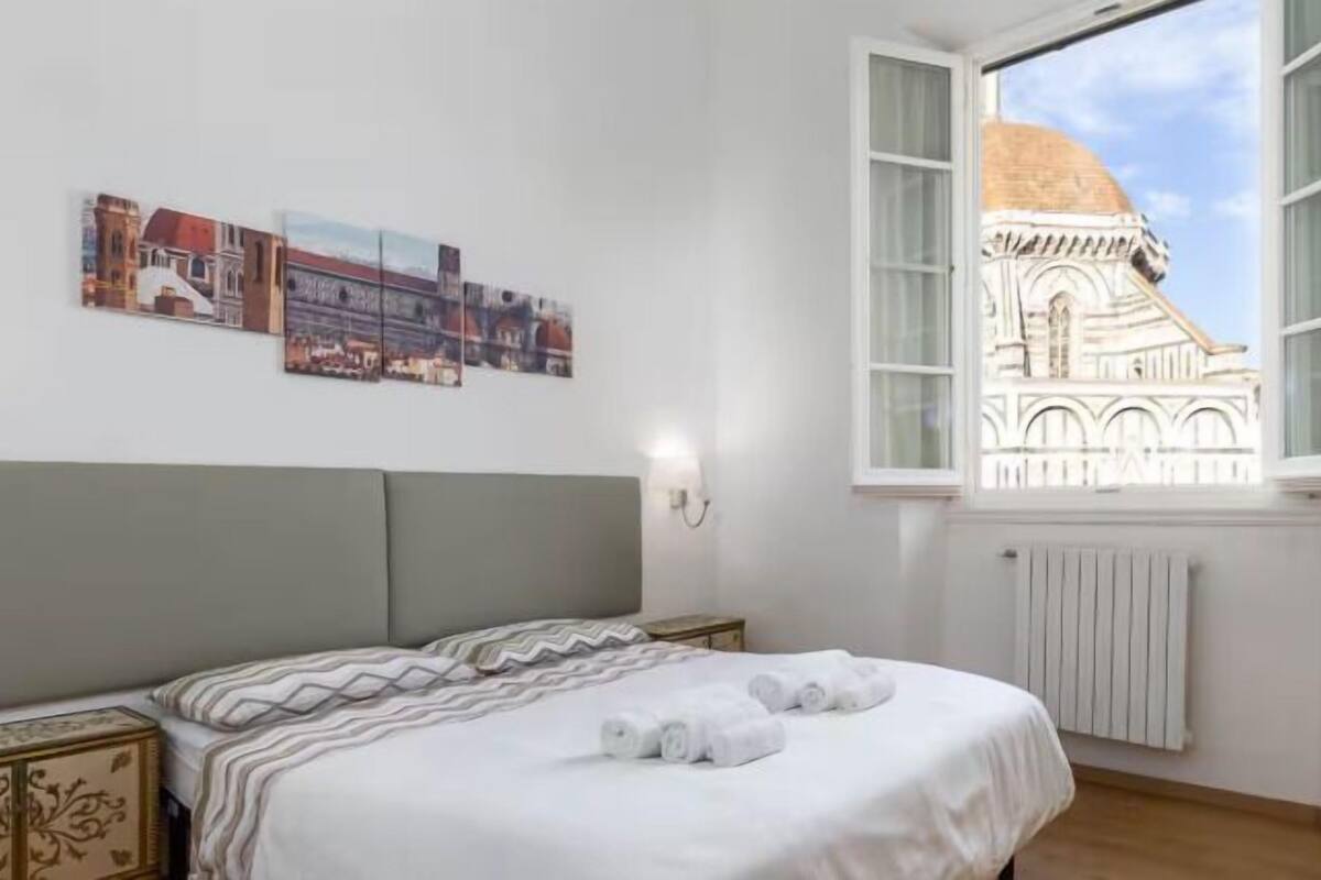 Una habitación de hotel con una cama, una mesita de noche, una lámpara de pared y una ventana con vistas a un edificio.
