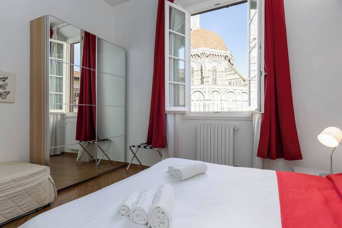 Habitación de hotel con cama, espejo, ventana con cortinas rojas, radiador y lámpara.