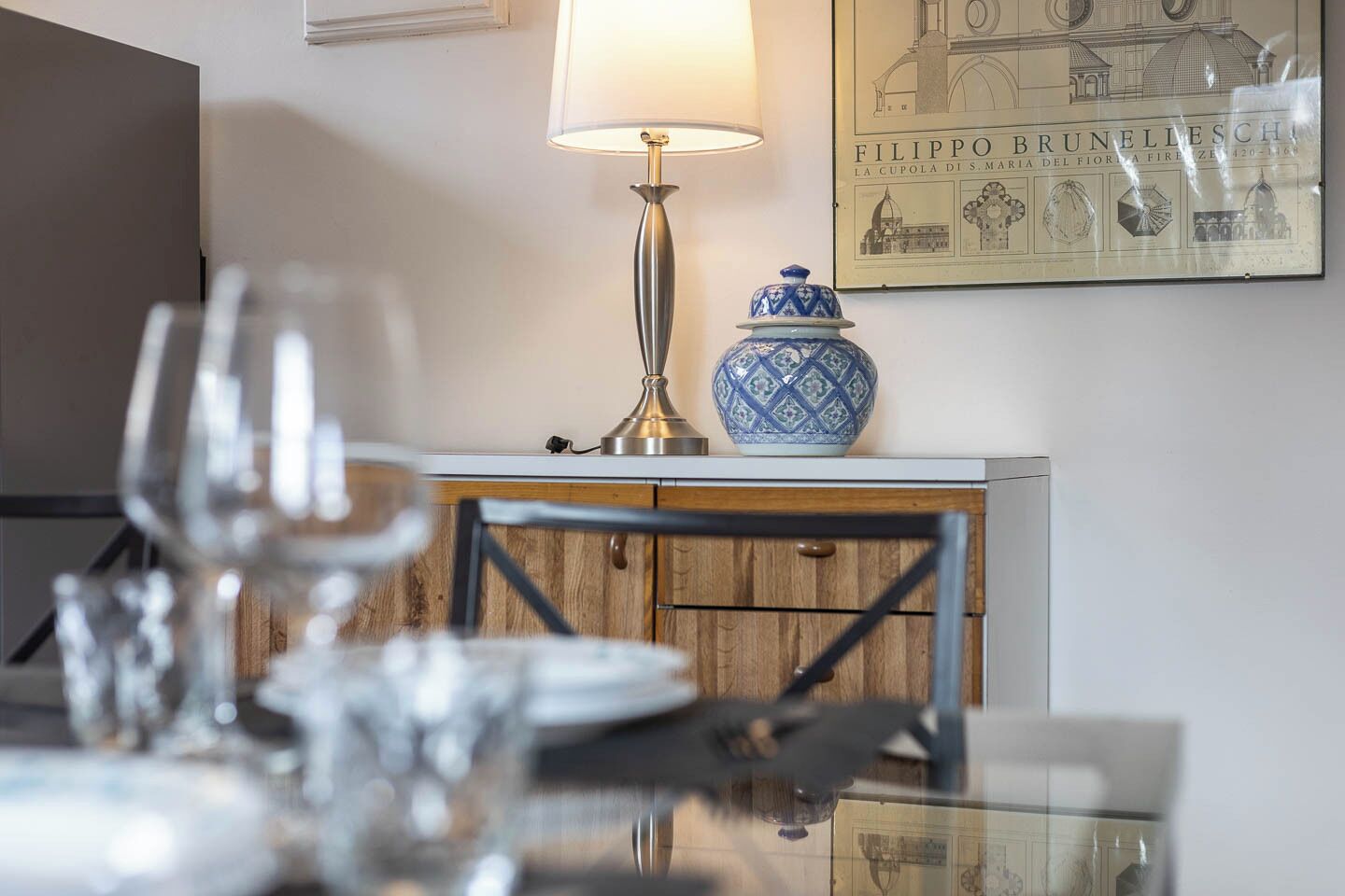 Una mesa de comedor con vasos y cubiertos, una lámpara, un jarrón decorativo y un cuadro en la pared.
