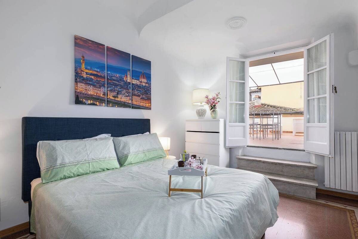 Un dormitorio con una cama grande, un cuadro de paisaje urbano, una ventana con una persiana abierta y vistas al balcón exterior.