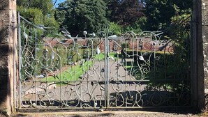 Un portail en fer forgé aux motifs complexes, installé dans un jardin bordé d’un mur de pierre et où l’on aperçoit des arbres au loin.