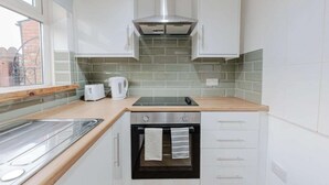 A compact kitchen with white cabinets, a stainless steel range hood, a built-in oven, a wooden countertop, and a window with a view of a brick building.
