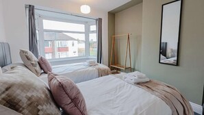 A hotel room with two beds, a window with curtains, a hanging wooden rack, and a framed mirror on the wall.