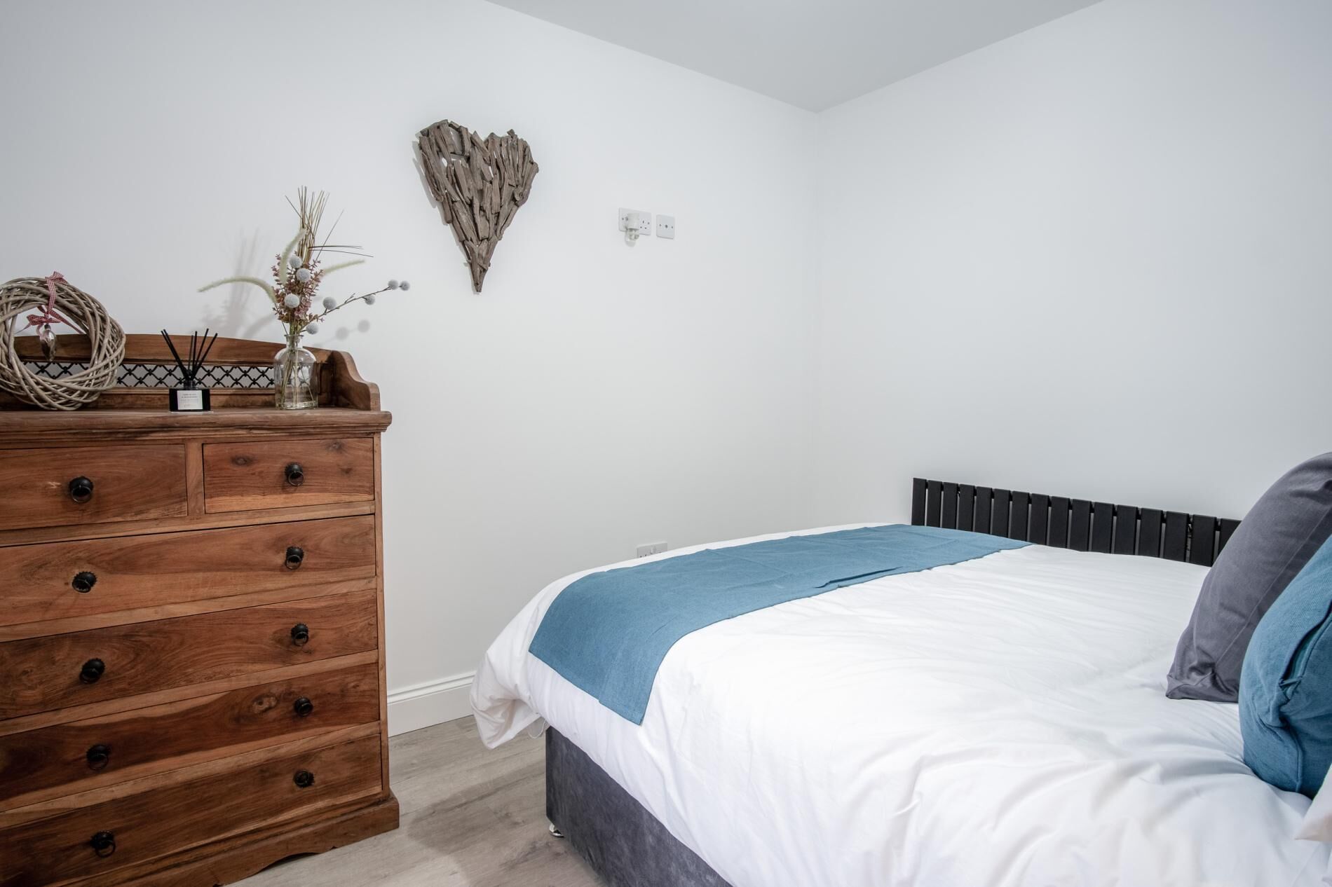 A bedroom with a wooden dresser, a bed with a blue and white bedspread, and a heart-shaped wall decoration.