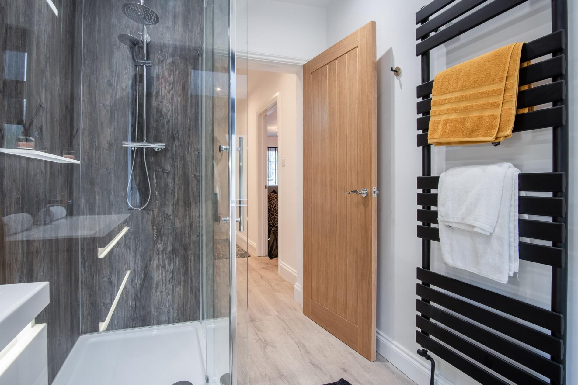 A modern bathroom with a glass-enclosed shower, a wooden door, and a towel rack with multiple towels.
