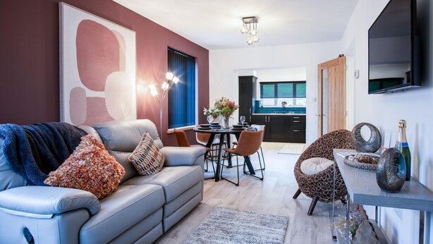 Ett modernt vardagsrum med en grå soffa, ett matbord med stolar, ett kök i bakgrunden och en tv som är monterad på väggen.