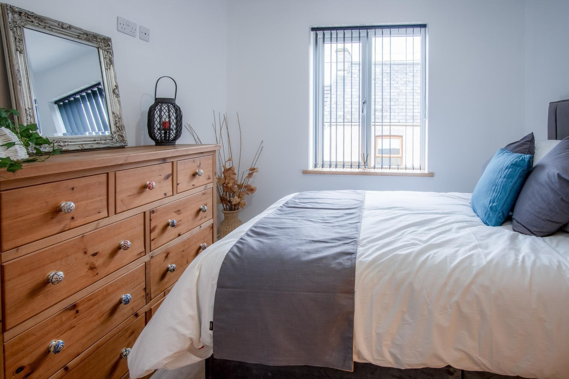 A bedroom with a wooden dresser, a mirror, a window with blinds, and a bed with pillows.