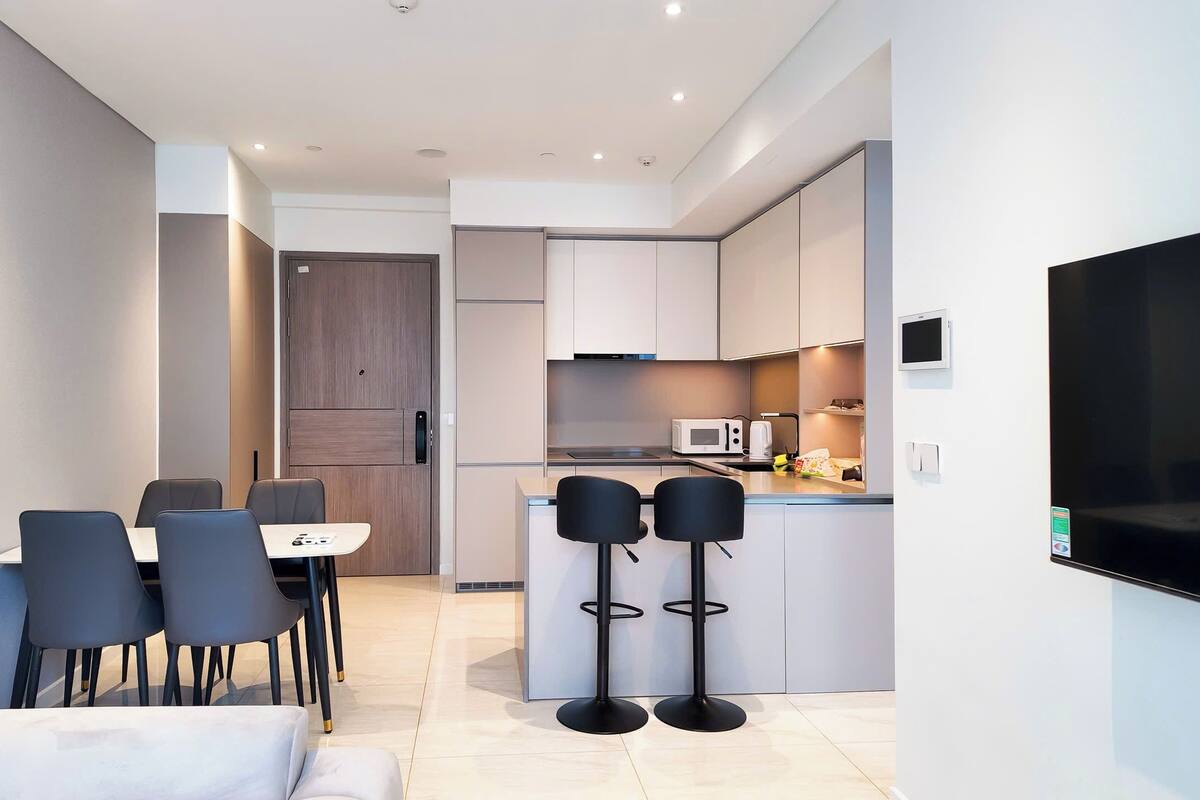 A modern kitchen with a microwave, stove, and a small island with bar stools. There is a dining area with a table and chairs, and a TV mounted on the wall.
