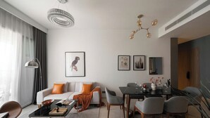 Ένα μοντέρνο σαλόνι με έναν καναπέ, ένα τραπεζάκι σαλονιού, ένα τραπέζι φαγητού και καρέκλες. Στον τοίχο υπάρχουν κορνιζάρισμενοι πίνακες και πάνω από το τραπέζι φαγητού κρέμεται ένα πολυέλαιος.