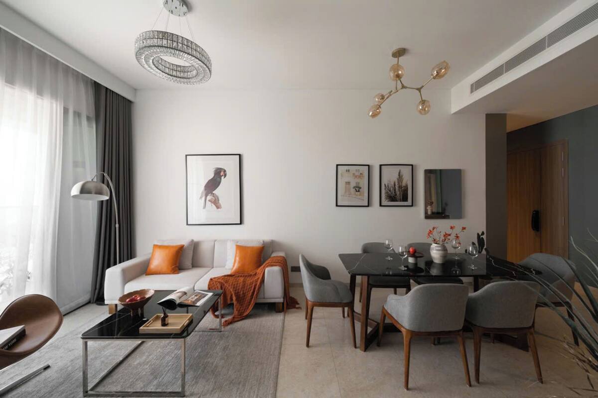 A modern living room with a sofa, coffee table, dining table, and chairs. There are framed pictures on the wall and a chandelier above the dining table.