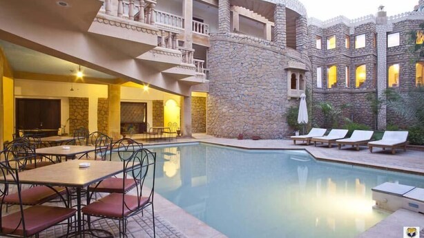 A poolside area with lounge chairs, a dining area with tables and chairs, and a stone building with balconies. - Deoki Niwas Palace (Jaisalmer)