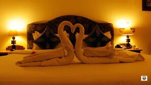 A hotel room with a bed featuring a heart-shaped towel arrangement, two bedside lamps, and a floral-patterned headboard. - Deoki Niwas Palace (Jaisalmer)