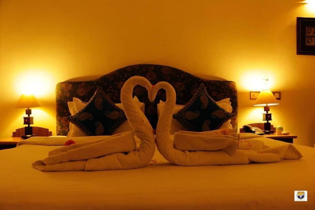 Une chambre d’hôtel avec un lit agrémenté d’une disposition de serviettes en forme de cœur, deux lampes de chevet et une tête de lit à motifs floraux.