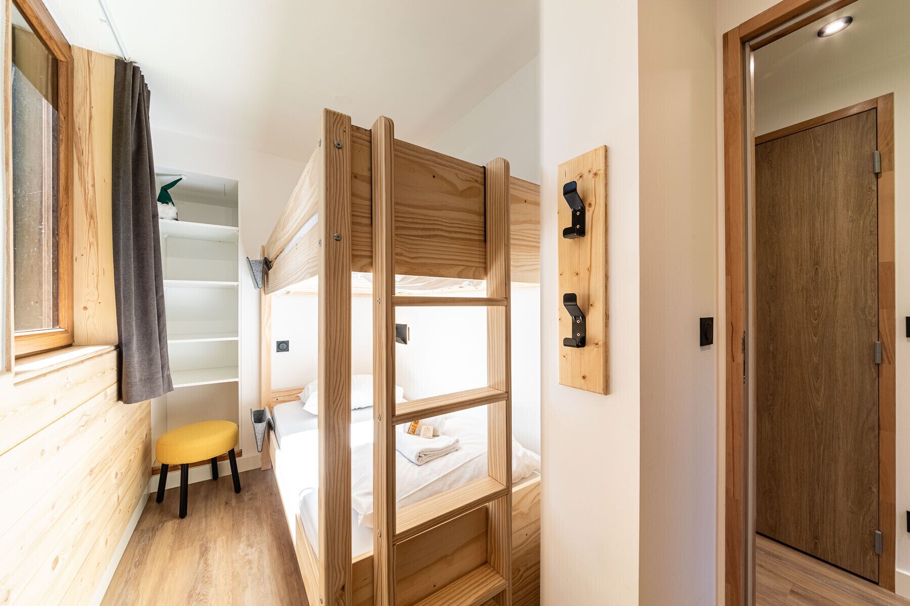 Une chambre avec un lit superposé, un plancher en bois, un petit tabouret et une porte.