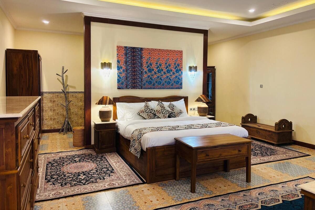 A well-lit bedroom with a large bed, two bedside tables, a wooden bench, and multiple rugs.