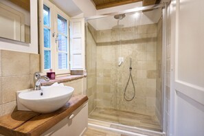 Un baño con lavamanos blanco, cabina de ducha de vidrio y ventana con contraventanas blancas.