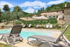 Un área junto a la piscina con sillones de descanso y sombrillas, rodeada de vegetación y edificios de piedra.