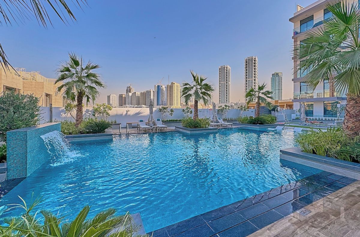 A swimming pool with palm trees and lounge chairs, surrounded by high-rise buildings.