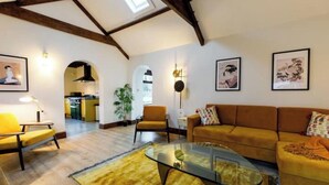 A spacious living room with a high ceiling, a glass coffee table, a sofa, and a chair. There are framed pictures on the walls and a plant in the corner. - The Granary - Rustic Retreats (Reynoldston)
