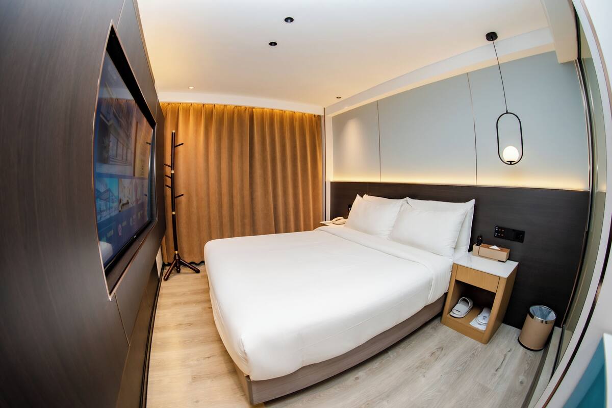 Een moderne hotelkamer met een groot bed, een televisie, een nachtkastje met lamp en een kleine prullenbak.