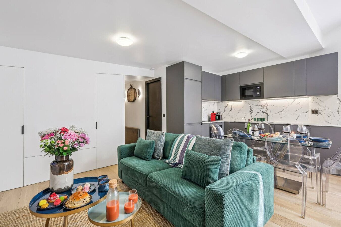 Ein modernes Wohnzimmer mit einem grünen Sofa, einem Esstisch mit klaren Stühlen und einer Küchenzeile mit grauen Schränken und einer Marmor-Spritzwand.